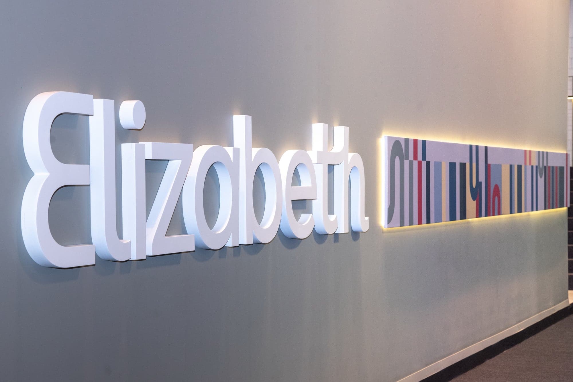 Logotipo Elizabeth em letras brancas iluminadas em relevo sobre parede clara, ao lado de um painel horizontal com listras verticais em várias cores, o grafismo do estande.