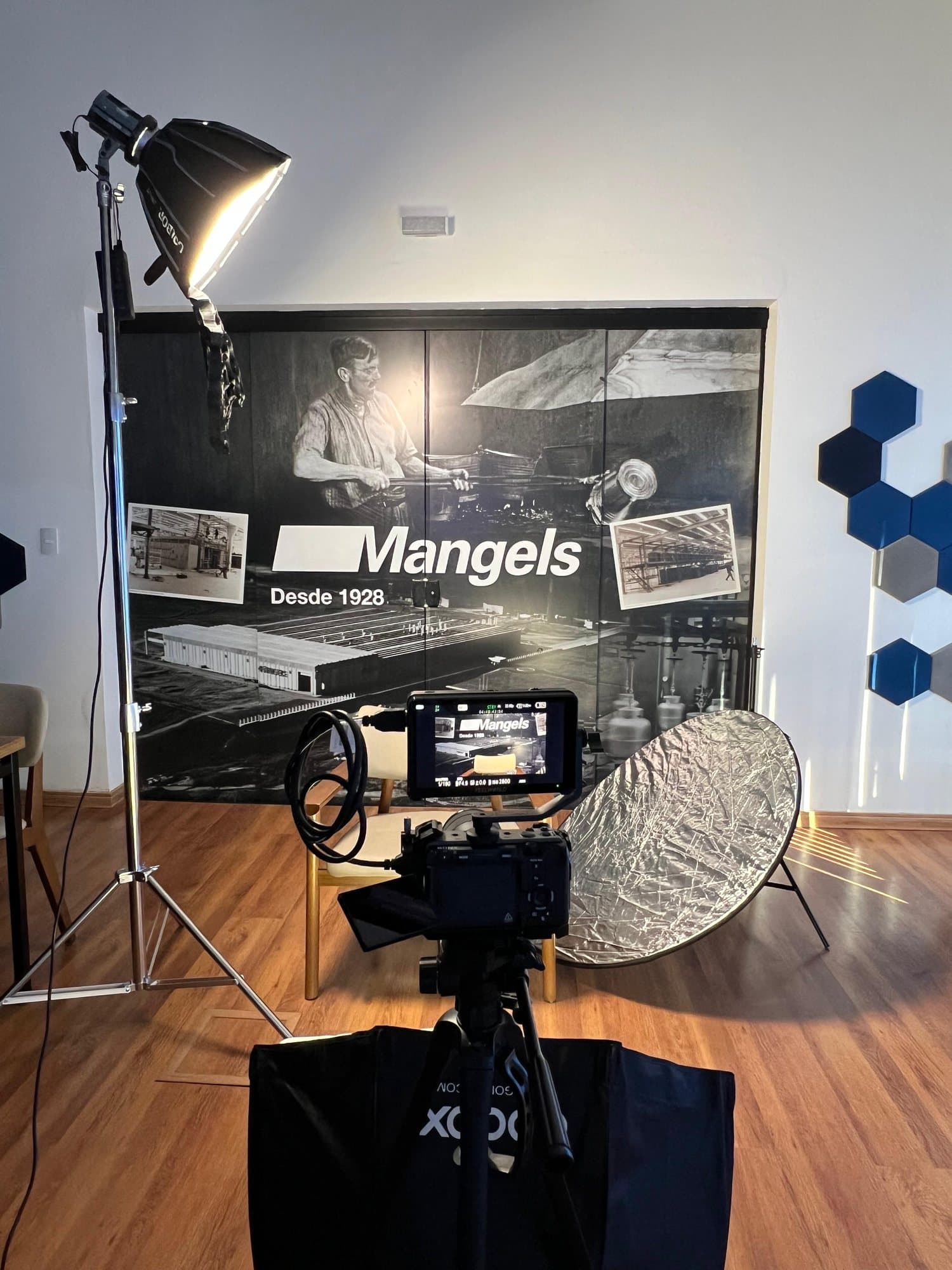 Gravação de um painel histórico da Mangels: fotos em preto e branco da fábrica sob a marca e a inscrição Desde 1928, iluminado por um refletor de estúdio, com a câmera em primeiro plano.