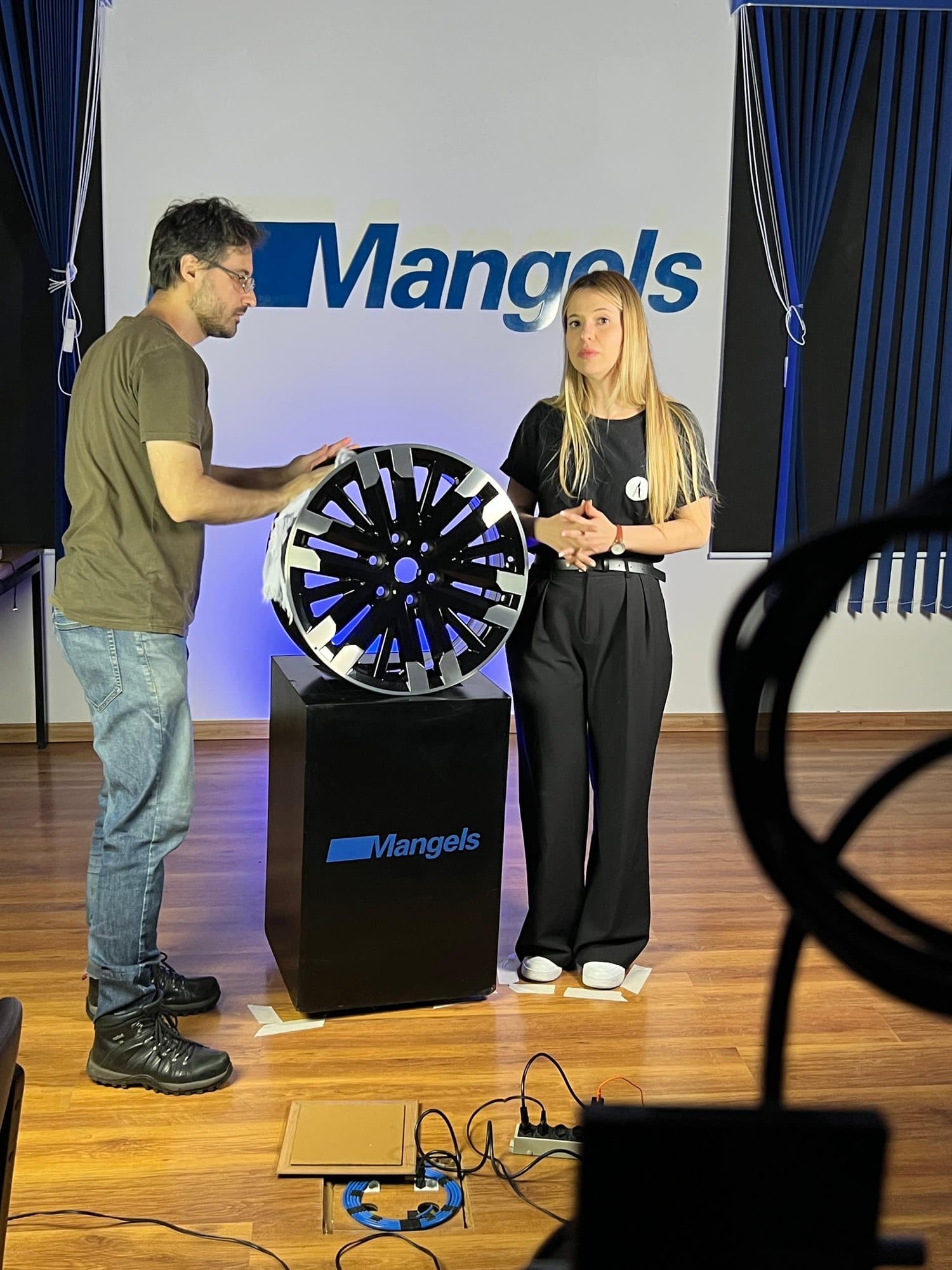 Gravação de episódio de série: duas pessoas apresentam uma roda de alumínio sobre um pedestal com a marca Mangels, diante de um painel azul com o logotipo da empresa.