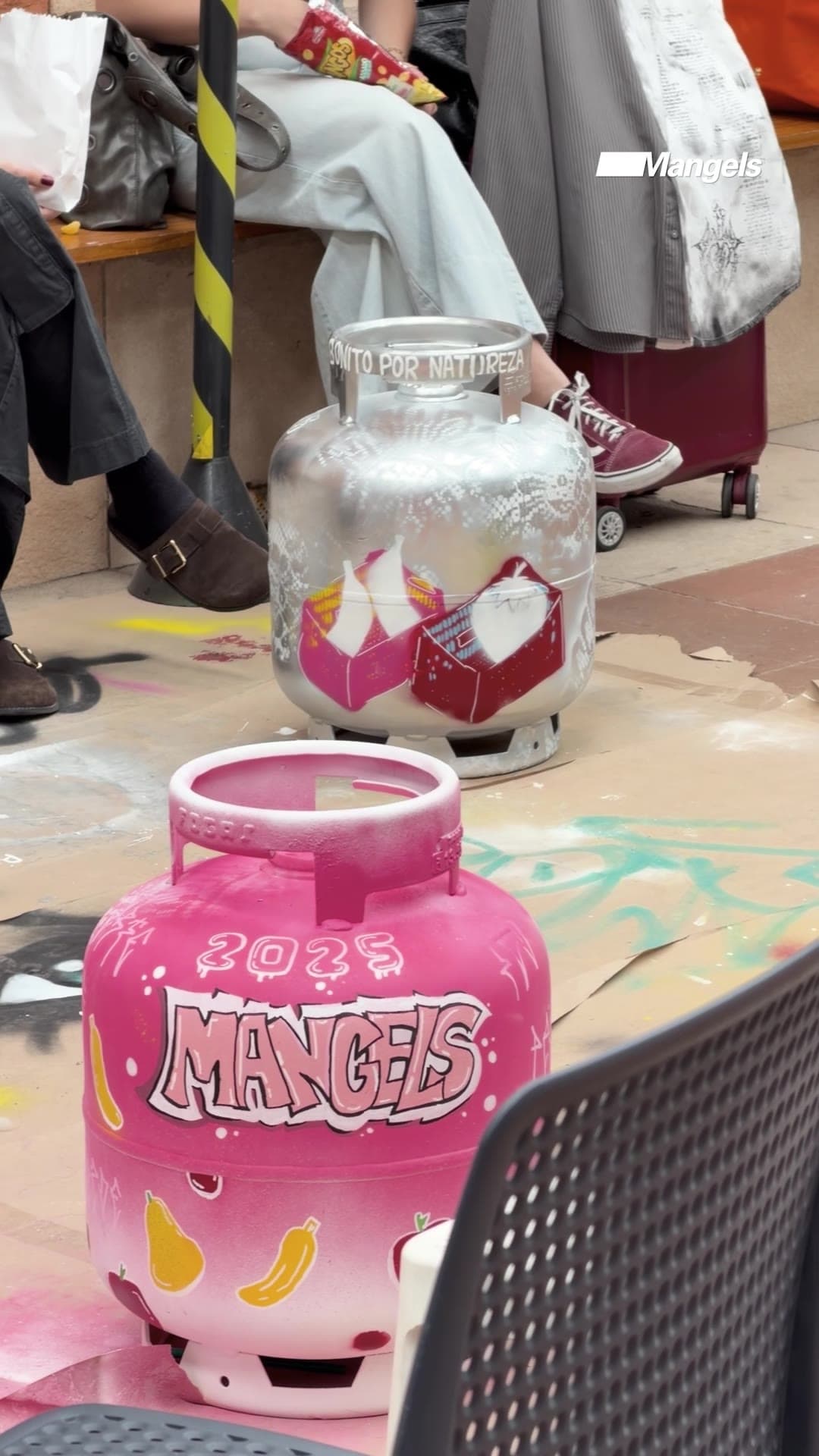 Botijão de gás pintado de rosa no Workshop Botijão Criativo do IED, com o nome Mangels em letreiro estilo grafite e o ano 2025, num ateliê com outros botijões pintados ao fundo.